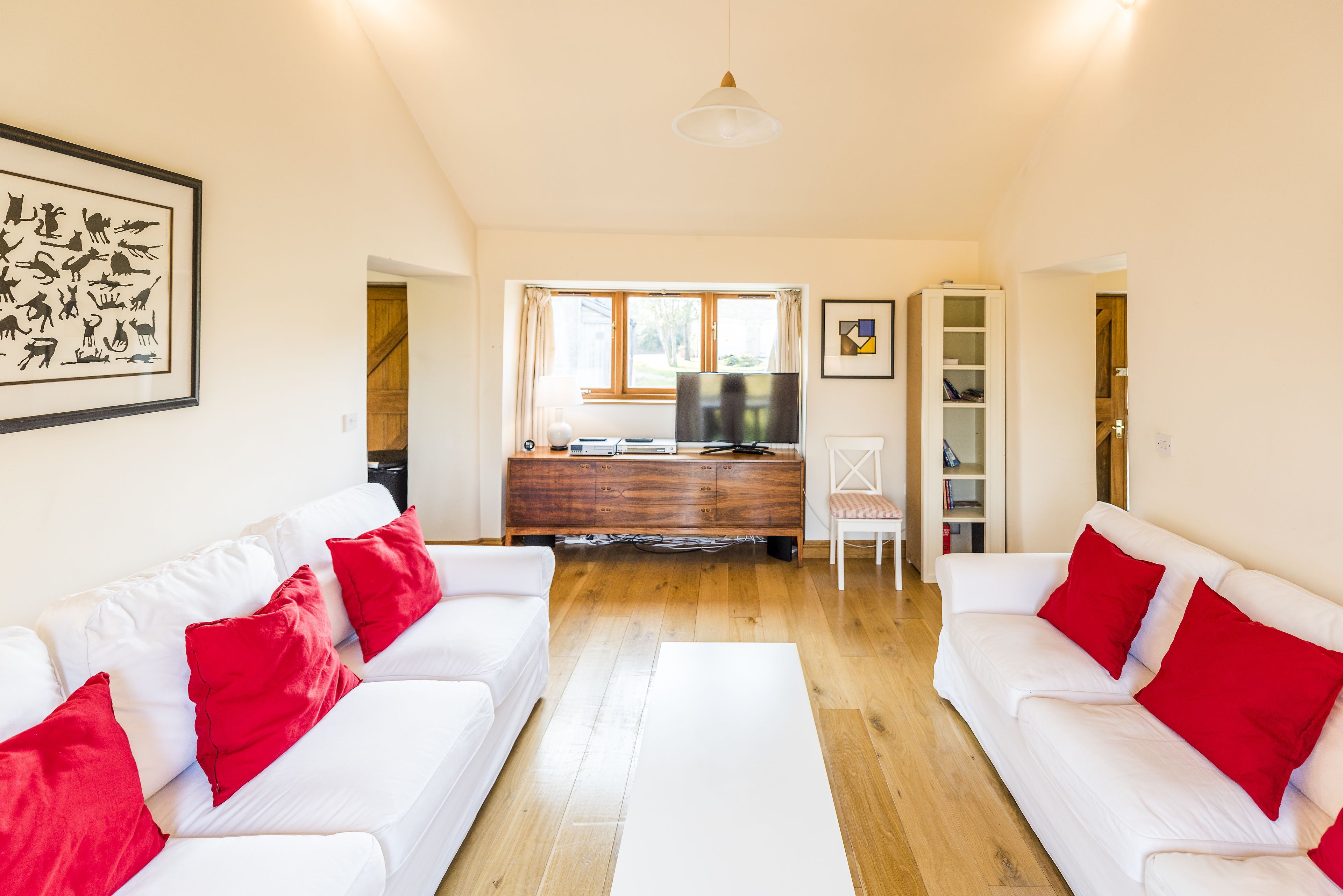 Un salón con sofá blanco, cojines rojos, un mueble de madera y televisión.