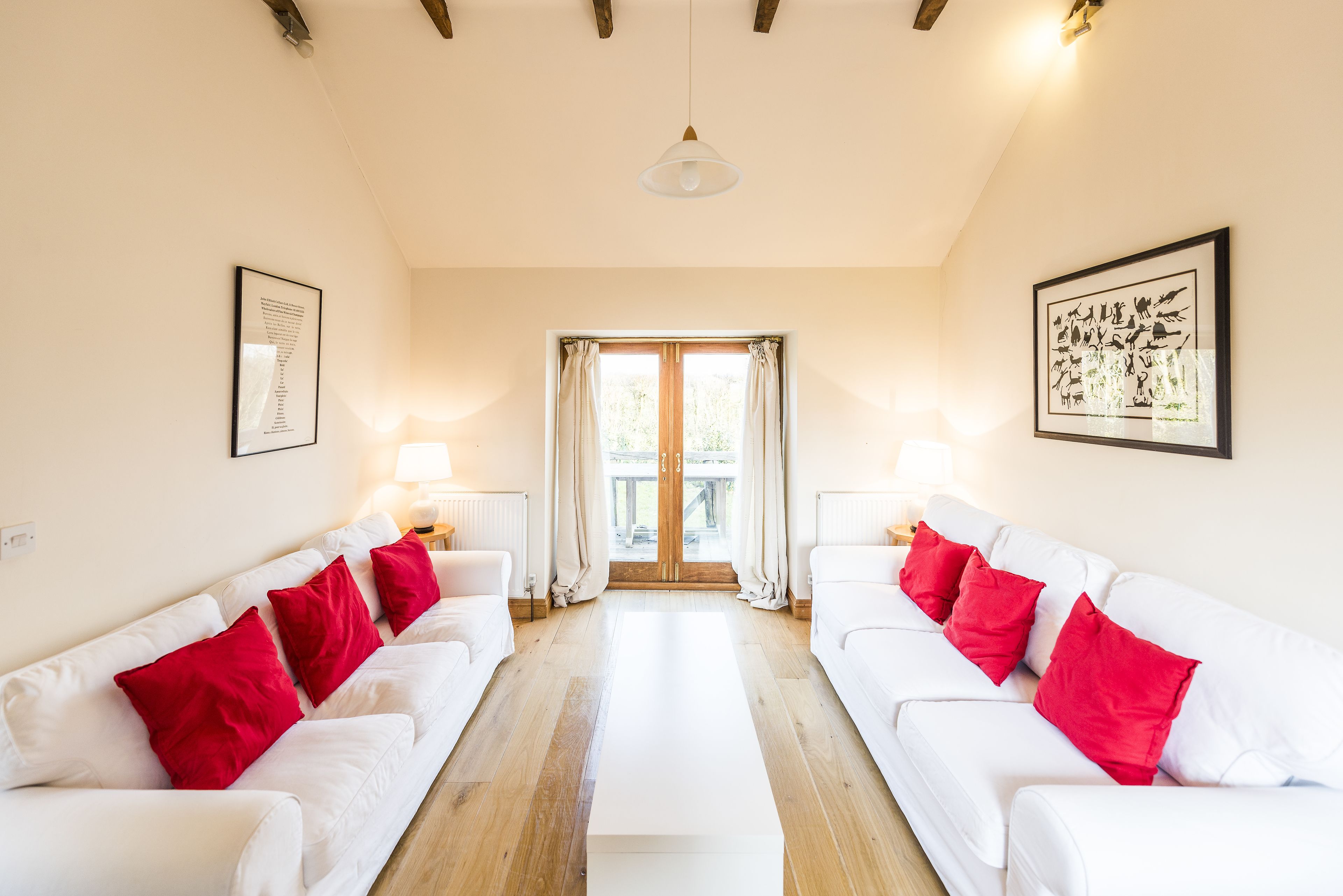 A spacious room with two white sofas and red cushions, a wooden floor, and a large window with curtains.