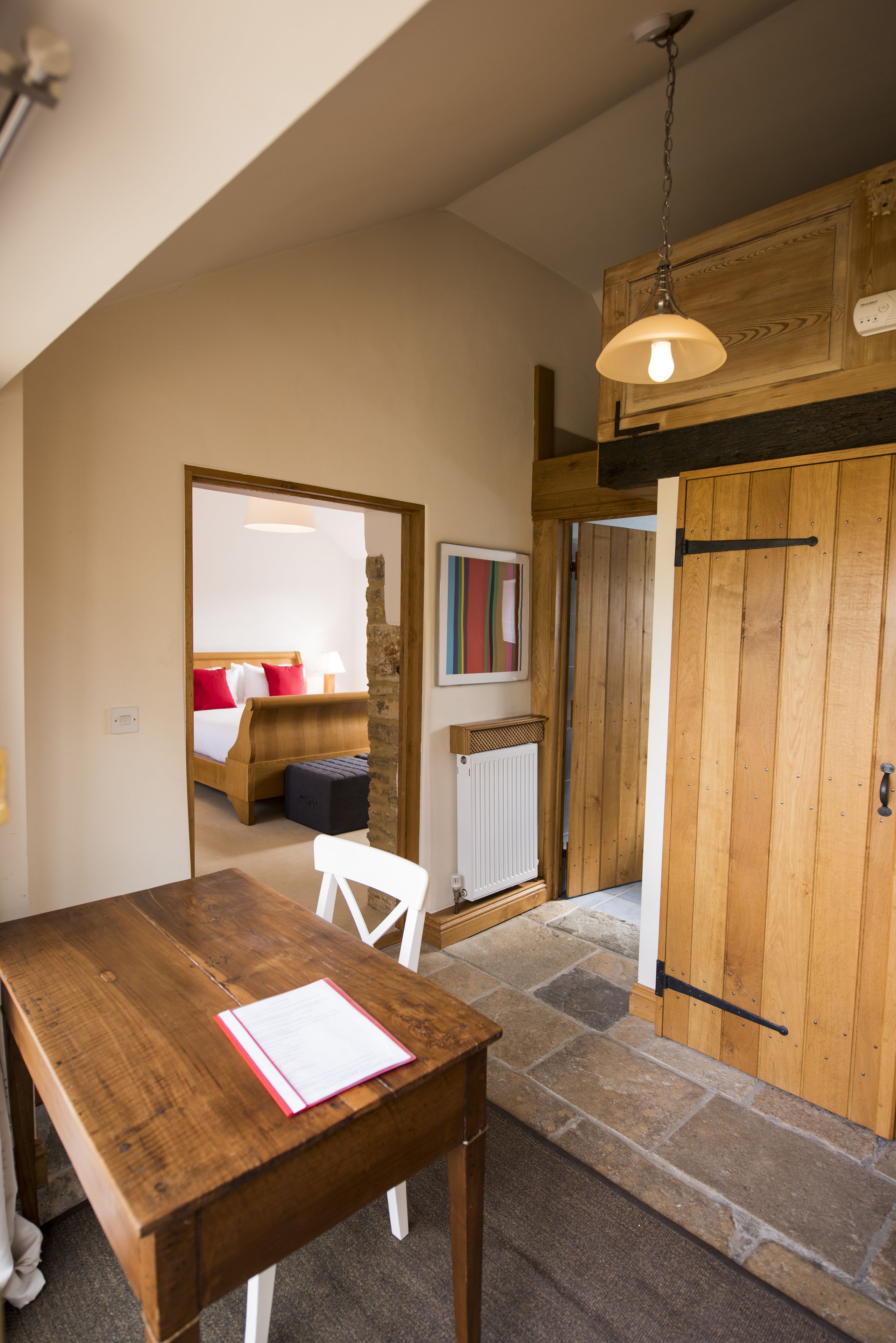 A wooden dining table with a chair and a book, a wooden door, a bed with red pillows, and a painting on the wall.