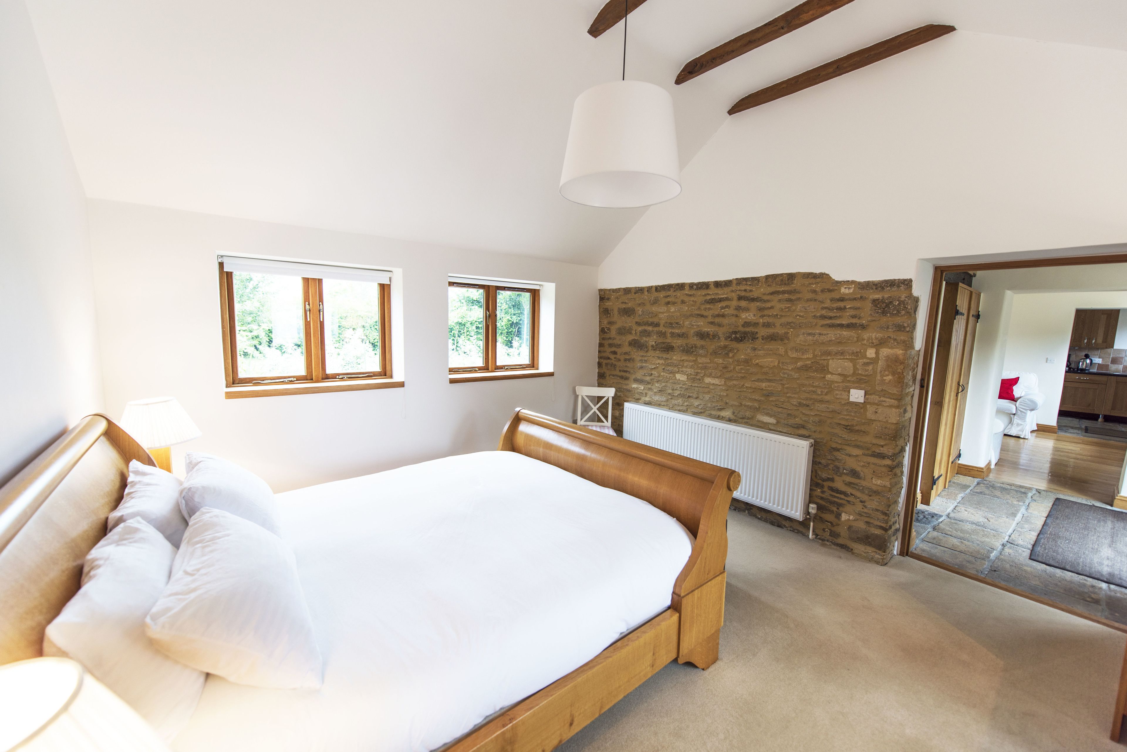 Een slaapkamer met een houten bed, twee ramen, een stenen muur en uitzicht op een andere kamer.