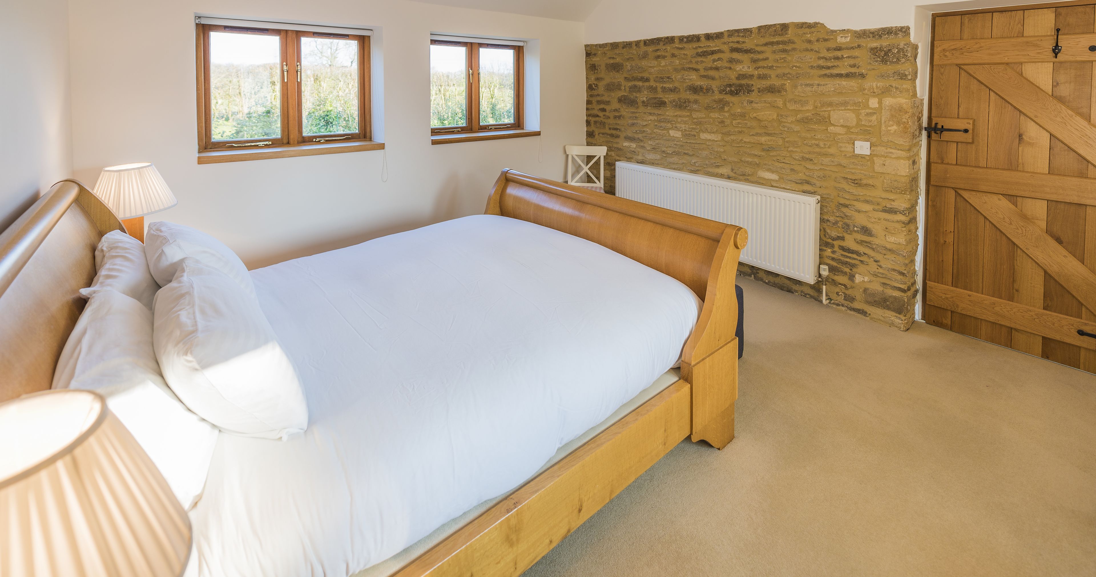 A bedroom with a wooden bed, two windows, a stone wall, a radiator, and a wooden door.