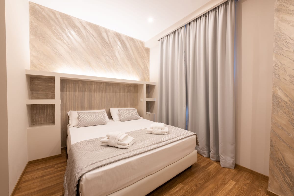 A modern hotel room with a large bed, wooden headboard, and a view of the city through sheer curtains.