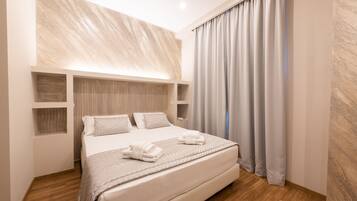 A modern hotel room with a large bed, wooden headboard, and a view of the city through sheer curtains.