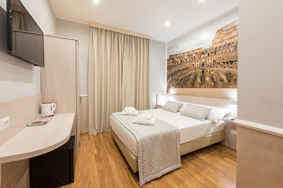 A hotel room with a bed, a desk, a television, and a mural of the Colosseum.