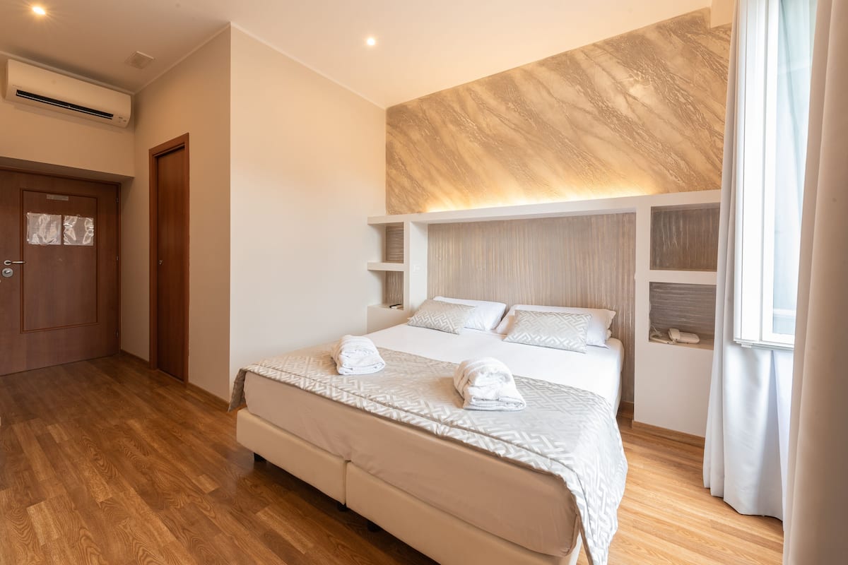 A modern hotel room with a large bed, wooden flooring, a built-in shelf, and a window with curtains.