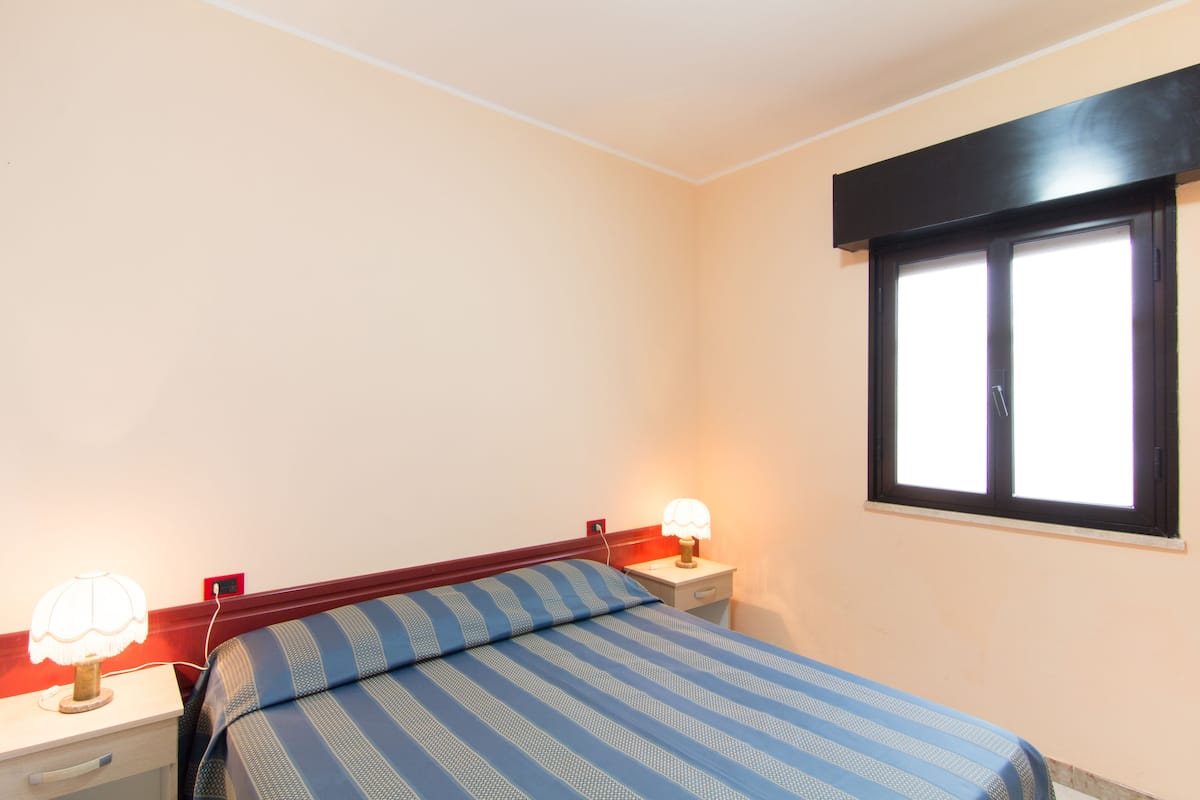 A bedroom with a bed, two bedside tables, a window, and a wall socket.