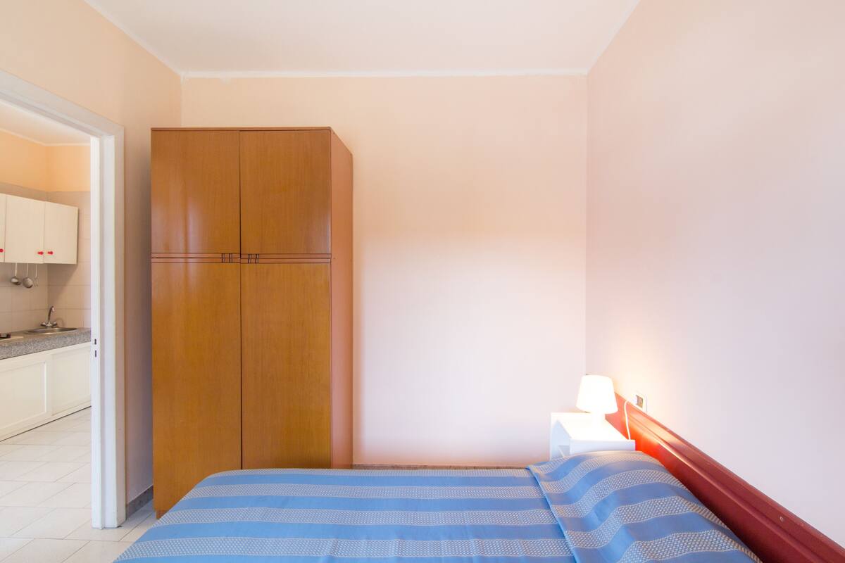 A bedroom with a bed, a wooden wardrobe, a bedside table with a lamp, and a view of a kitchen area.