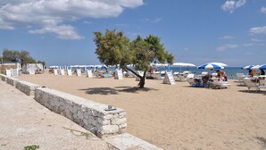 A beach with white lounge chairs and blue umbrellas, a stone wall, and a tree in the center. - Racar Hotel & Resort (Lecce)