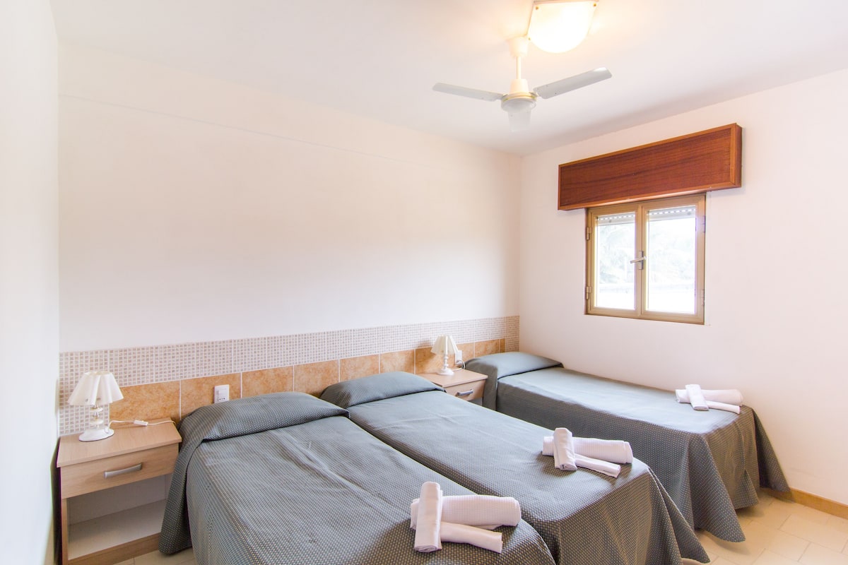 A hotel room with two single beds, a nightstand with a lamp, a ceiling fan, and a window with blinds.