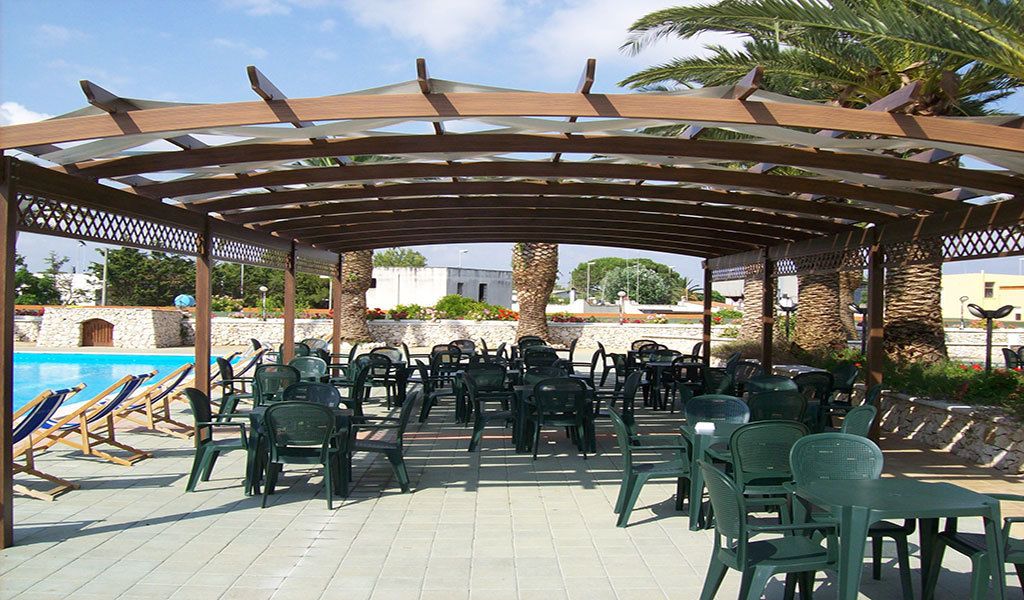Ein überdachter Außenbereich mit Stühlen und Tischen, im Hintergrund ein Pool und Palmen.