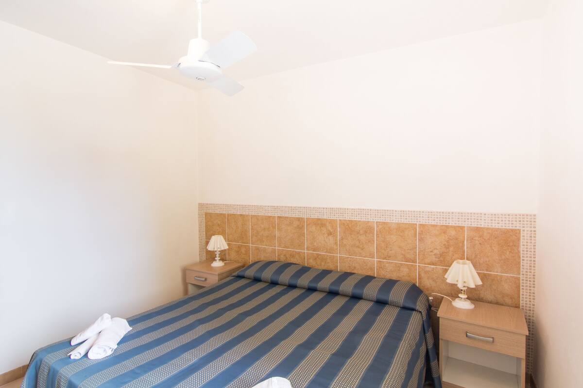 A hotel room with a bed, two bedside tables, a ceiling fan, and a wall-mounted light fixture.
