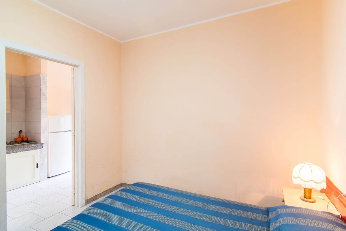 A bedroom with a bed, a nightstand with a lamp, a bathroom with a sink and mirror, and a door leading to another room.
