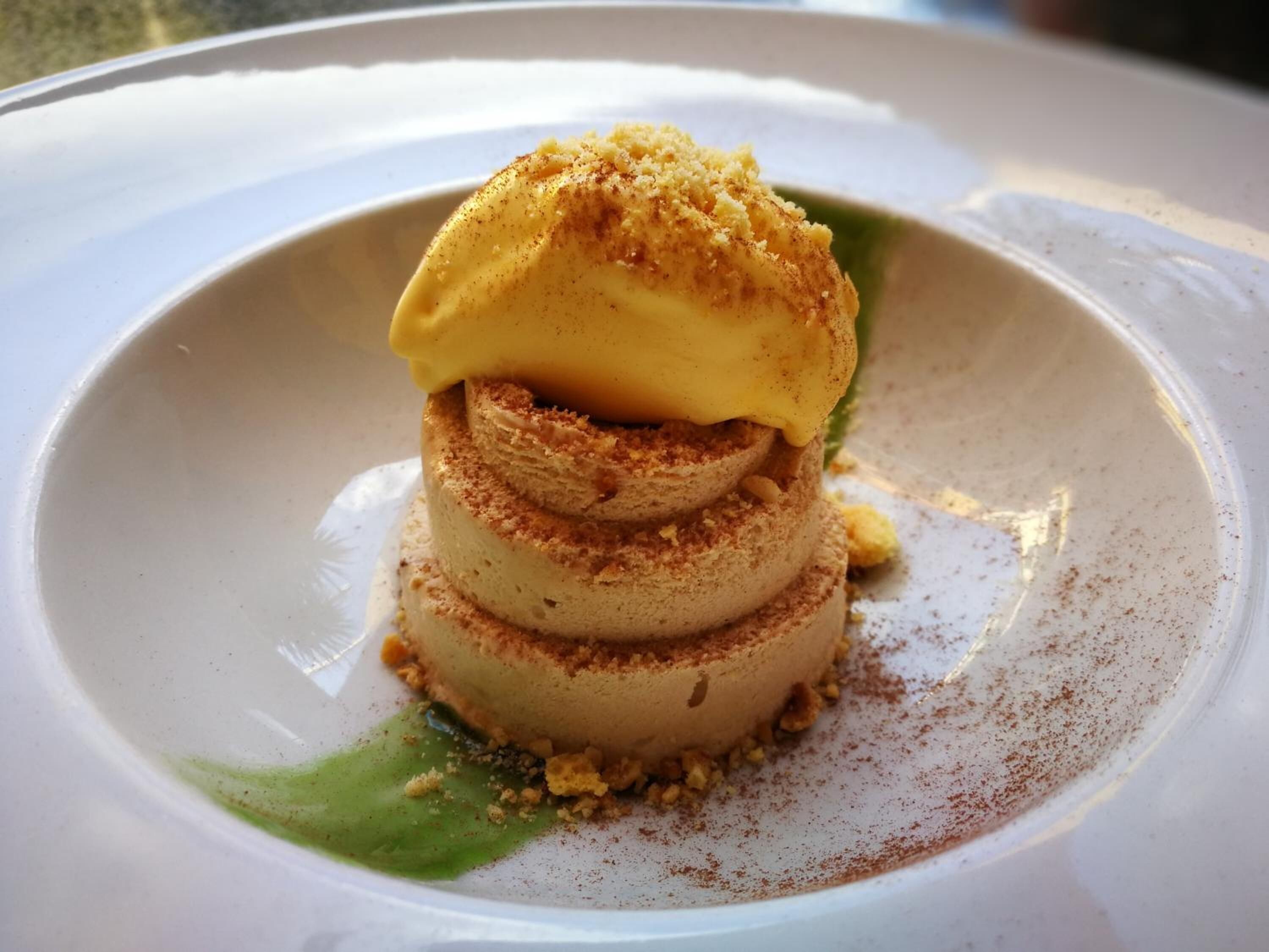 A dessert with a layered structure, topped with a golden-brown crumble, served on a white plate with a green garnish.