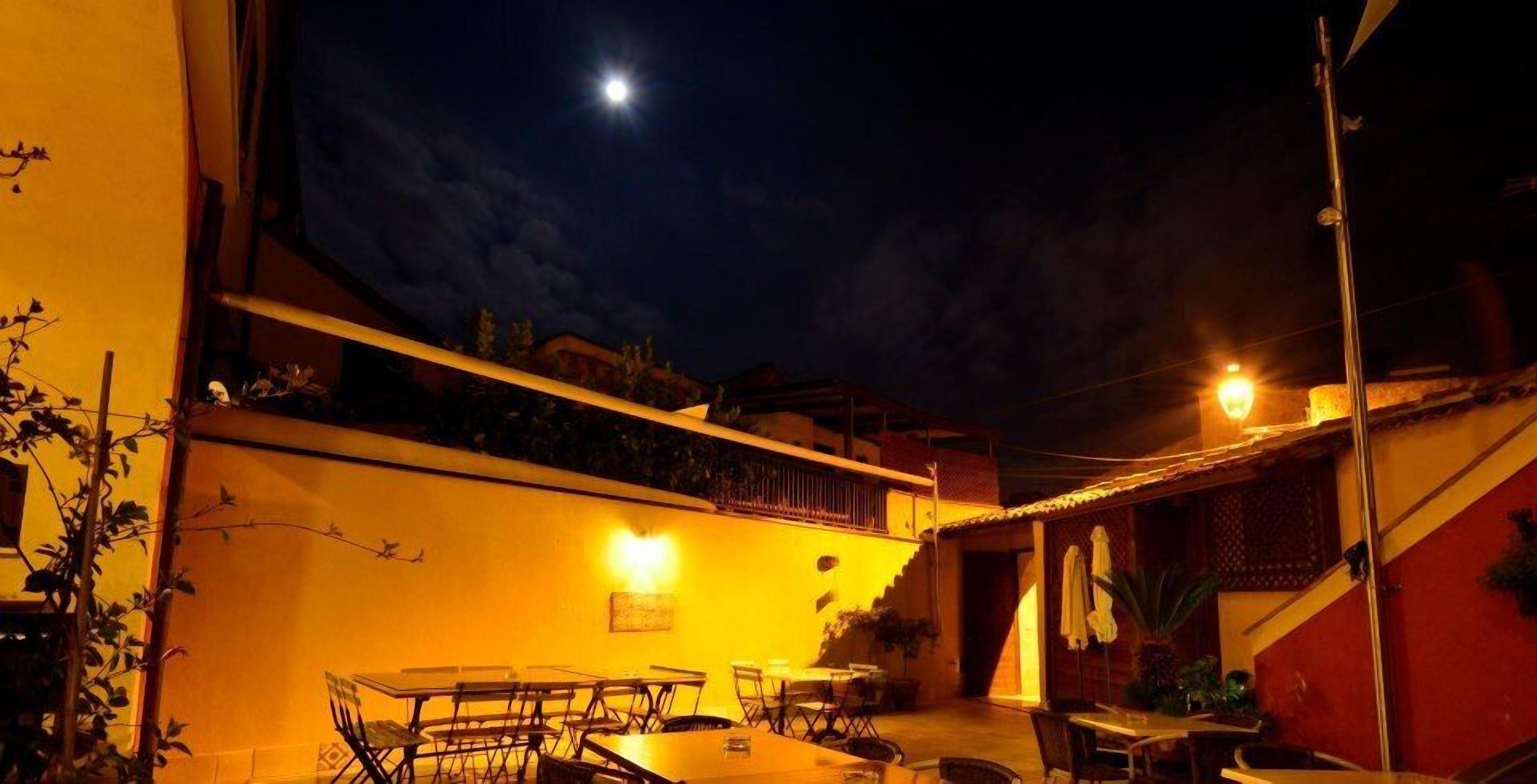 Un área de comedor al aire libre con mesas y sillas, una escalera y vistas al cielo nocturno con una luna visible.