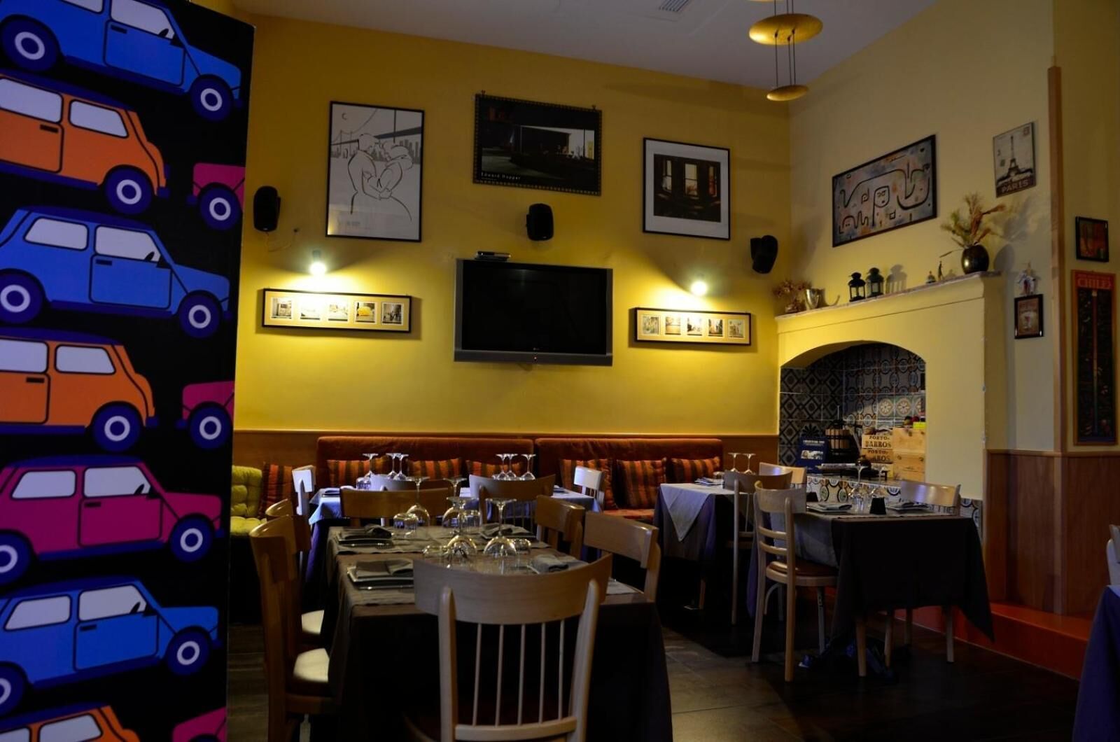 A restaurant with a yellow wall, multiple framed pictures, and a television. Tables are set with wine glasses and plates.