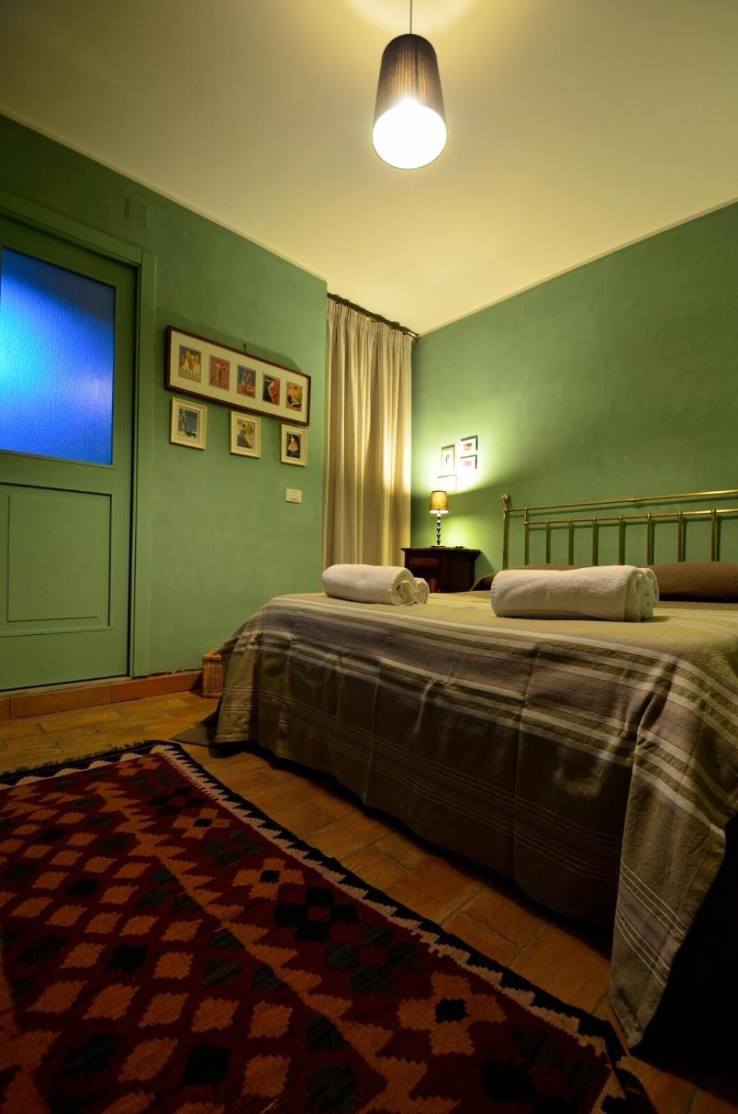 Une chambre à coucher avec un lit, une table de chevet, une lampe, une fenêtre avec des rideaux et des tableaux encadrés accrochés au mur.