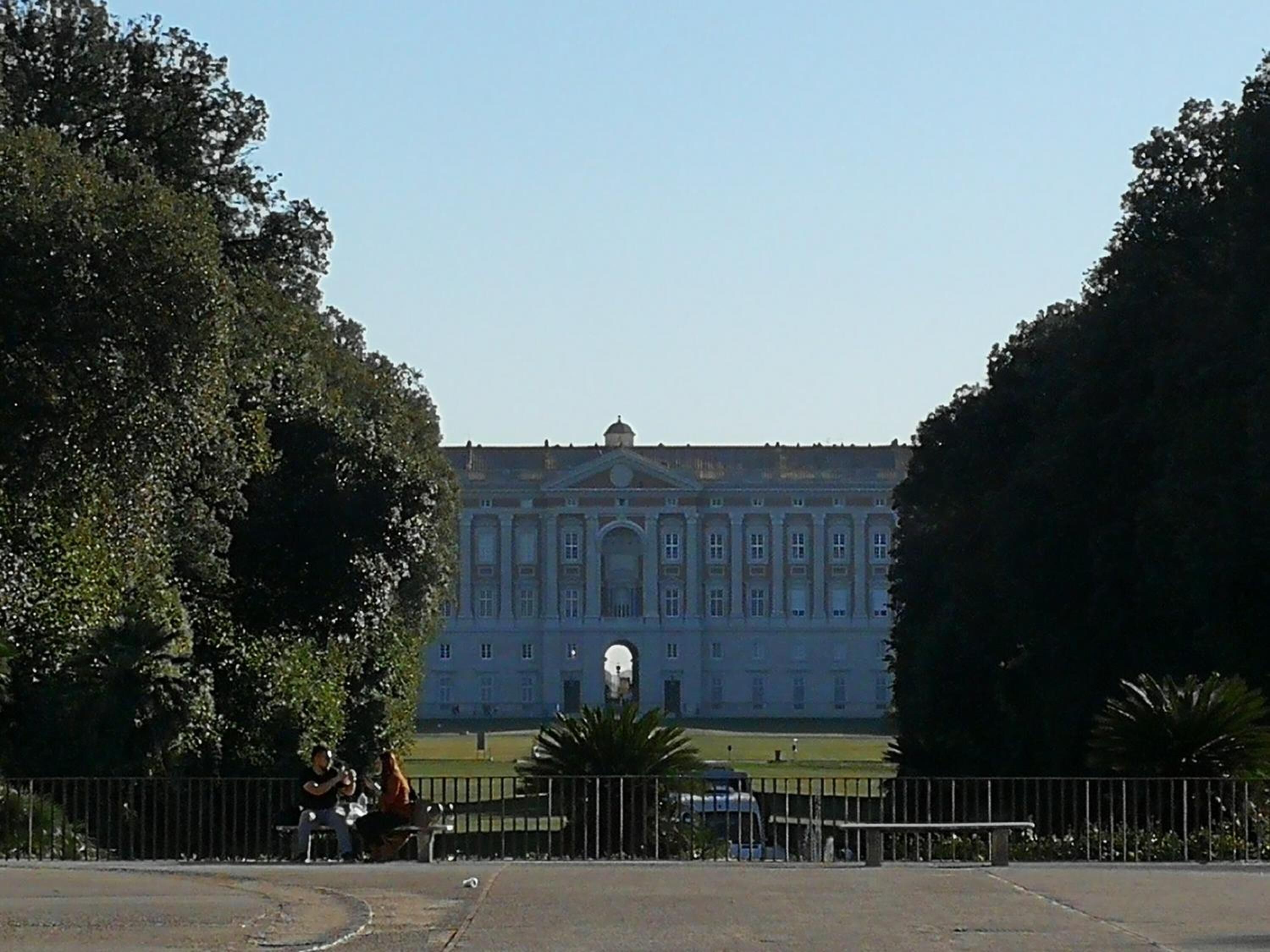A large, classical building with symmetrical architecture, likely a museum or historical site, surrounded by trees and a fenced area with benches.