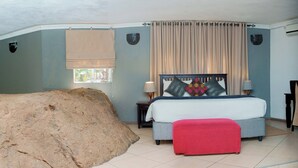 A bedroom with a large rock, a bed with pillows, a nightstand, and a window with curtains. - Sethule Lodge (Bulawayo)