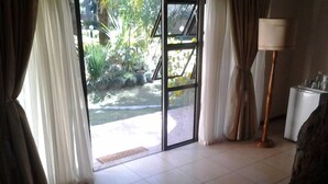 A room with a large sliding glass door leading to a garden area, a floor lamp, and a small white table. - Sethule Lodge (Bulawayo)