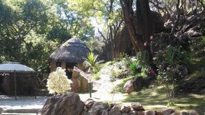 A rustic thatched-roof hut surrounded by a garden with rocks, plants, and a small white umbrella. - Sethule Lodge (Bulawayo)