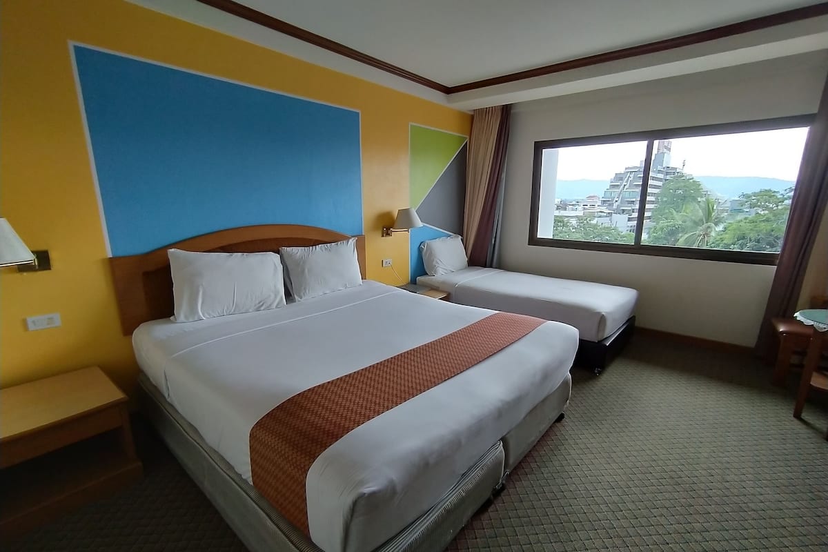 A hotel room with a large bed, two single beds, a window with a view, and a small table.