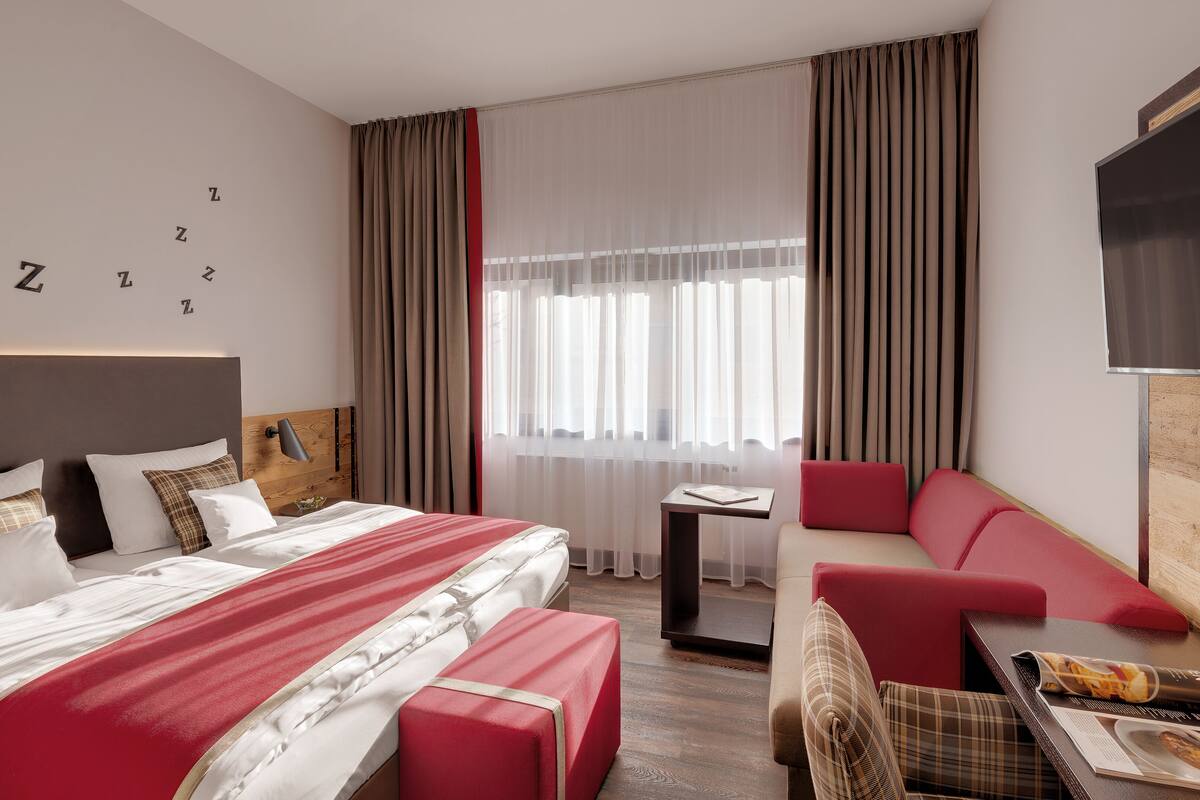 A hotel room with a large bed, a red sofa, a small table with a magazine, and a TV on the wall.