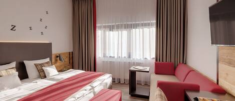 A hotel room with a large bed, a red sofa, a small table with a magazine, and a TV on the wall.