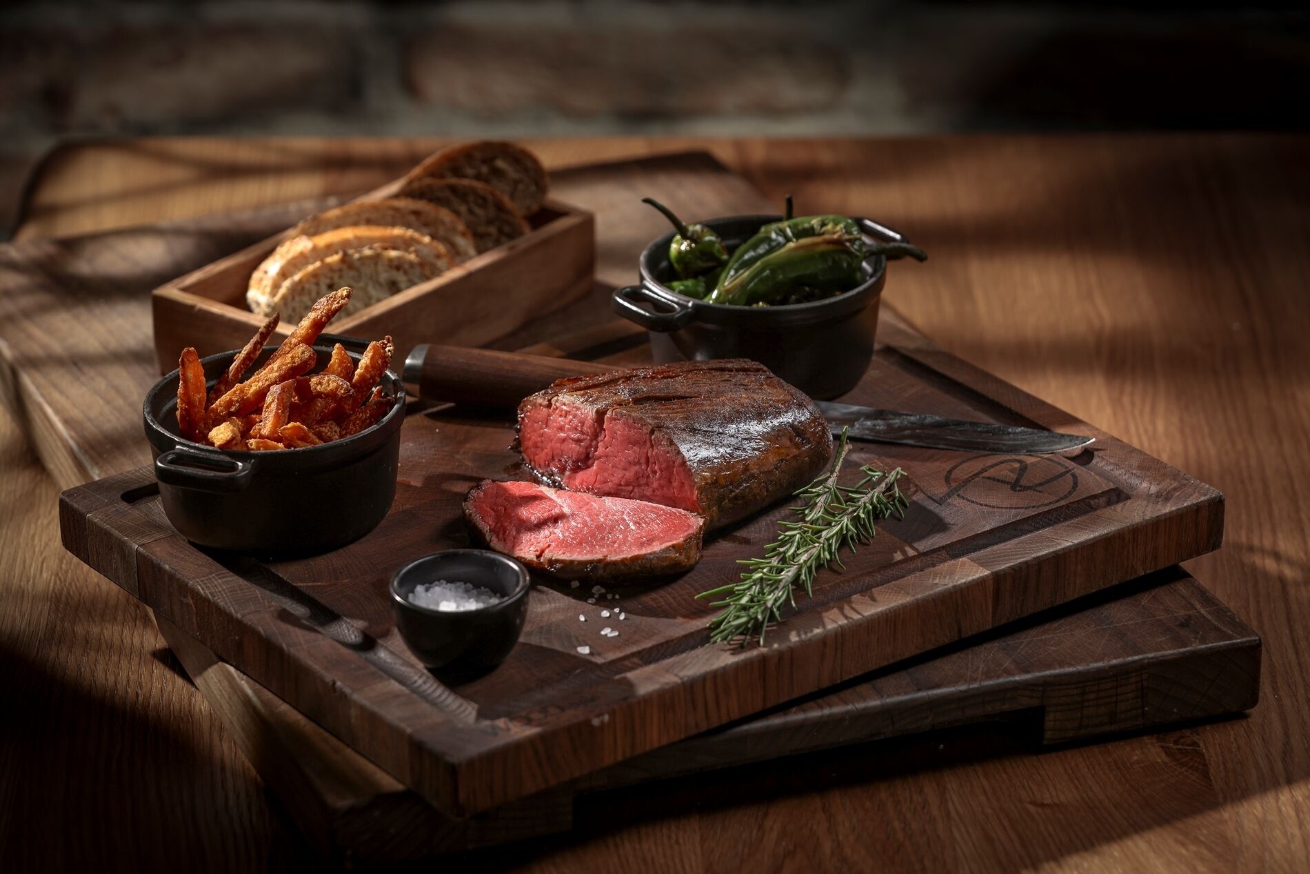 Een houten snijplank met een steak, brood, ingelegde pepers en een schaal zout.