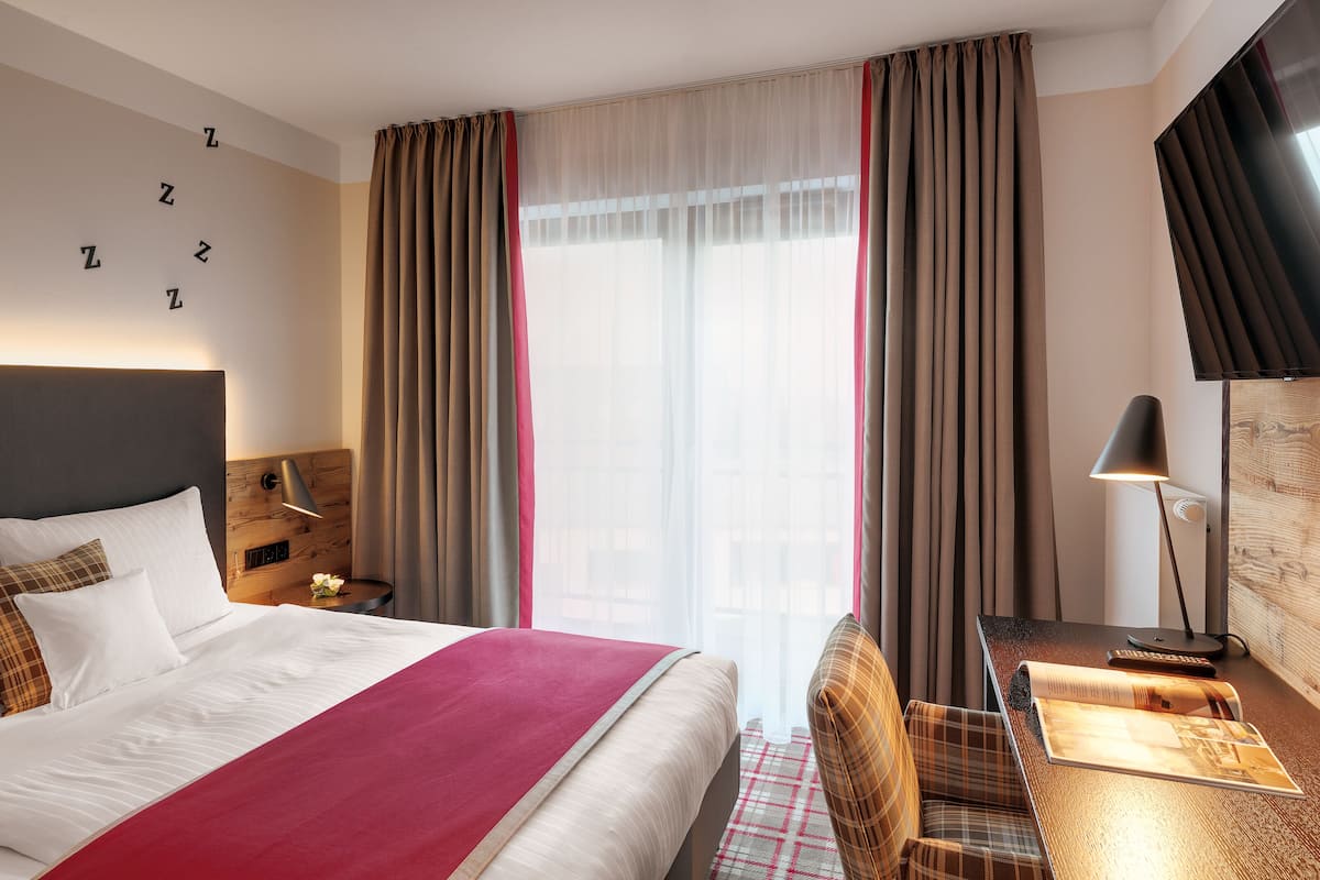 A hotel room with a large bed, a desk with a lamp, and a view of the city through sheer curtains.