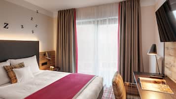 Une chambre d’hôtel avec un grand lit, un bureau avec une lampe et une vue sur la ville à travers de fines rideaux.