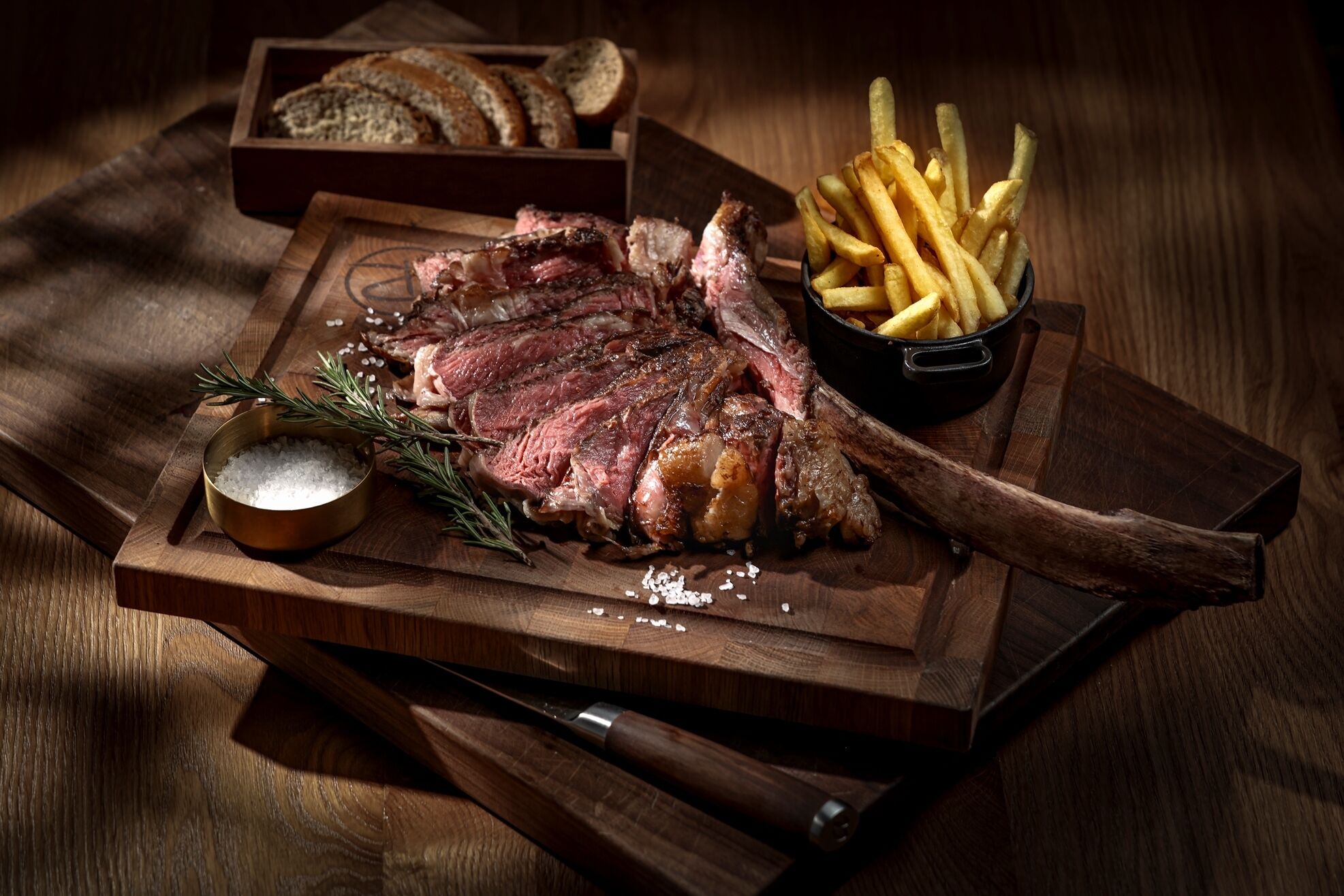 Un plato de carne con papas fritas sobre una tabla de madera, un cuenco de sal y un cuchillo.