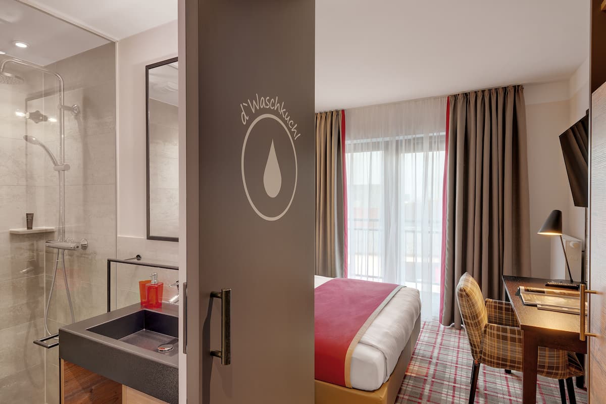 A modern hotel room with a shower, a bed, a desk, and a view of the outdoors.
