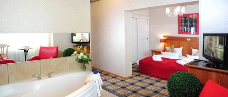Habitación de hotel con una bañera grande, una cama con ropa de cama roja, un televisor y una mesita con un jarrón de flores.