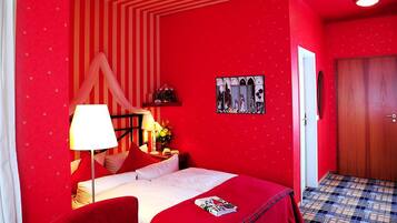 A red-themed hotel room with a bed, a nightstand, a lamp, and a painting on the wall.