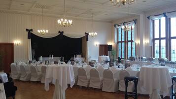 Une salle de banquet avec de nombreuses tables rondes dressées pour le repas, des lustres et de grandes fenêtres.