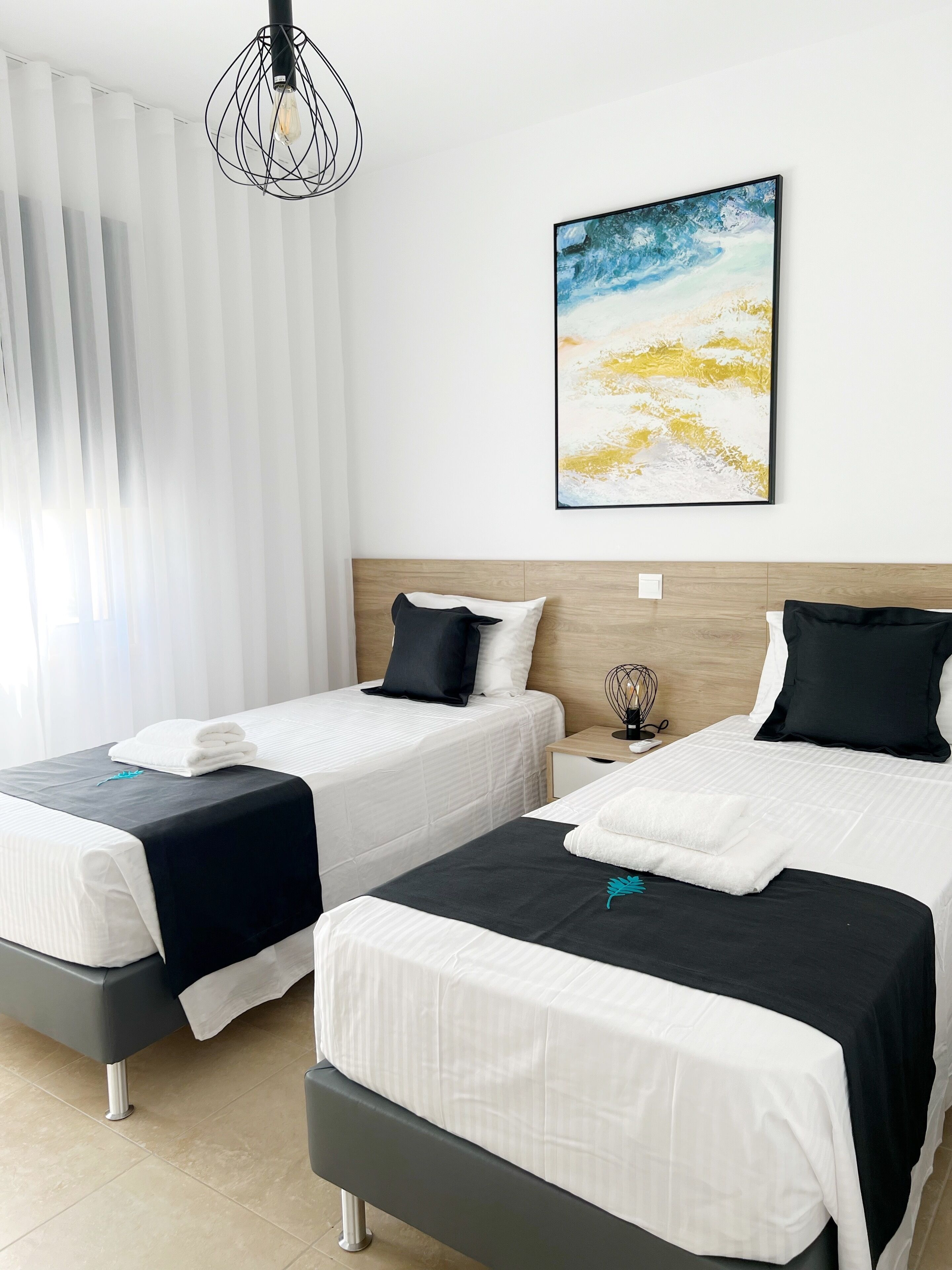 A hotel room with two beds, white bedding, a wooden headboard, a painting on the wall, and a modern light fixture.
