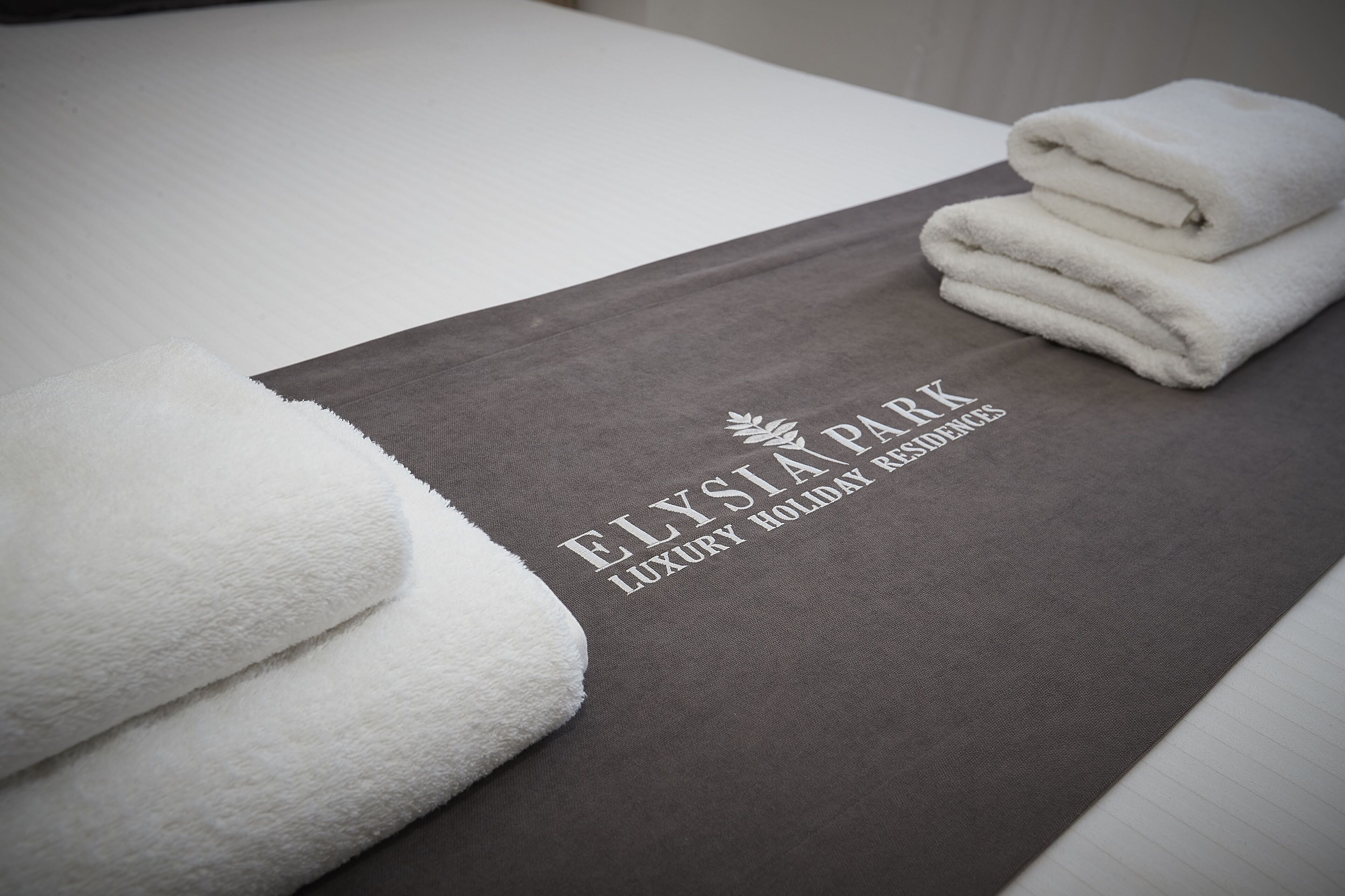 A neatly folded white towel and a brown placemat with the text "ELYSIA PARK LUXURY HOLIDAY RESORTS" on a bed.