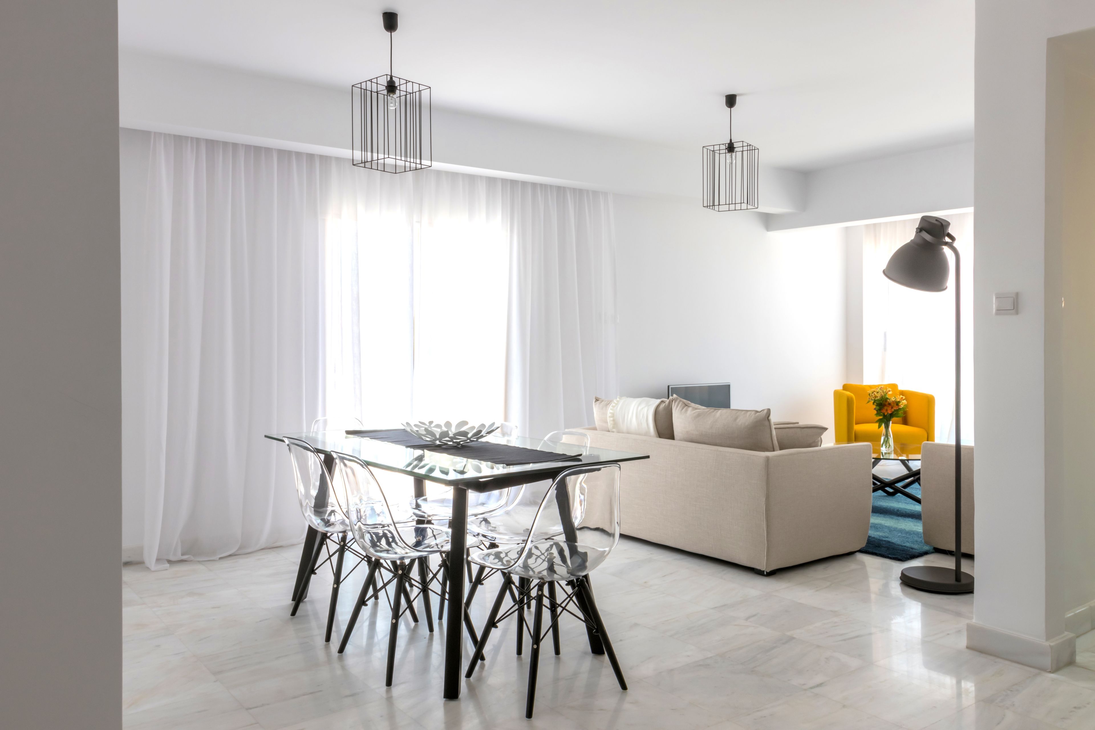 A modern living room with a glass dining table, clear chairs, a beige sofa, and a yellow armchair.