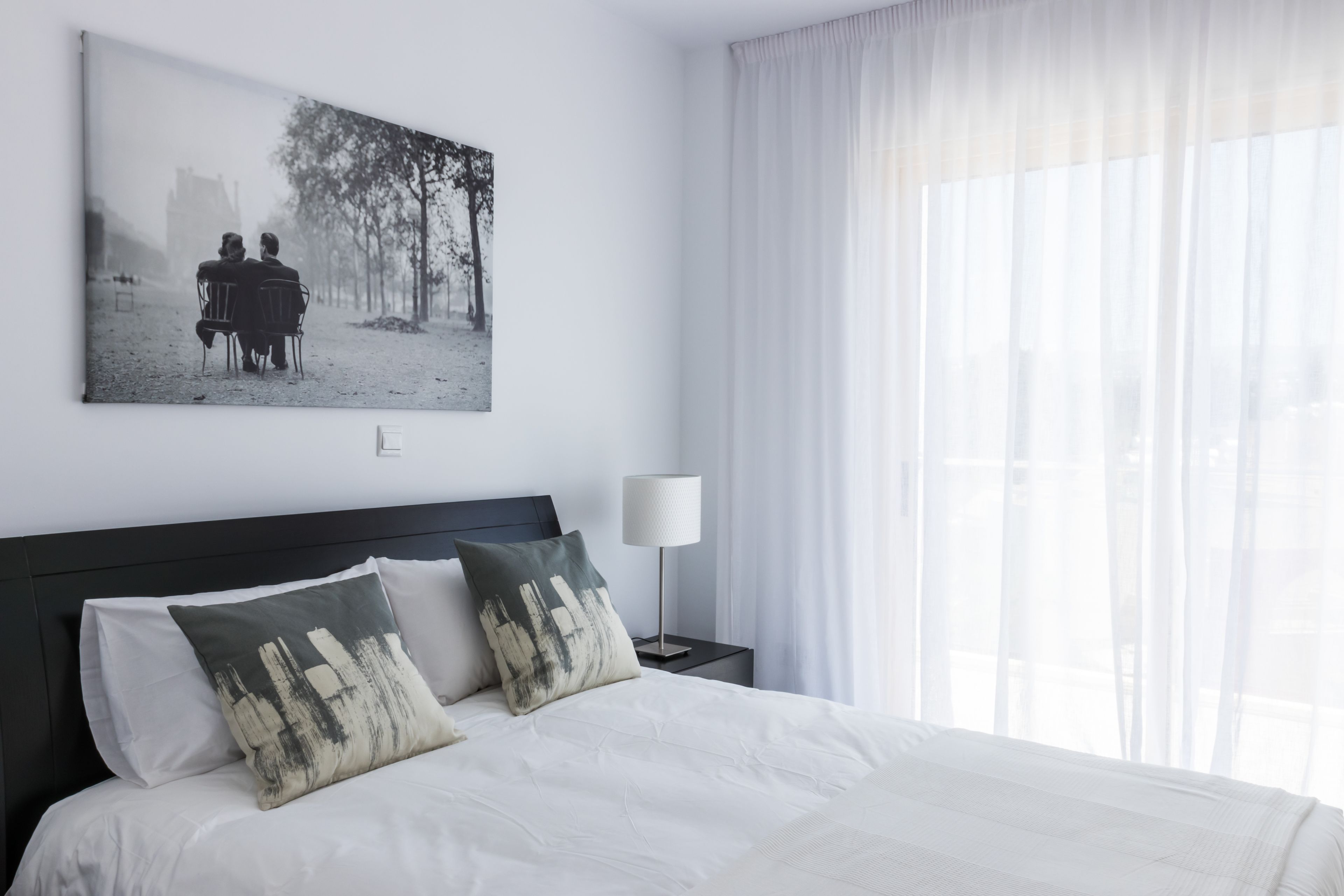 Una cama bien hecha con ropa blanca y dos almohadones decorativos, una mesita de noche con una lámpara, y una fotografía grande enmarcada en la pared.