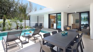 Un área de comedor exterior moderna con piscina, sillones de descanso y vistas a la cocina.