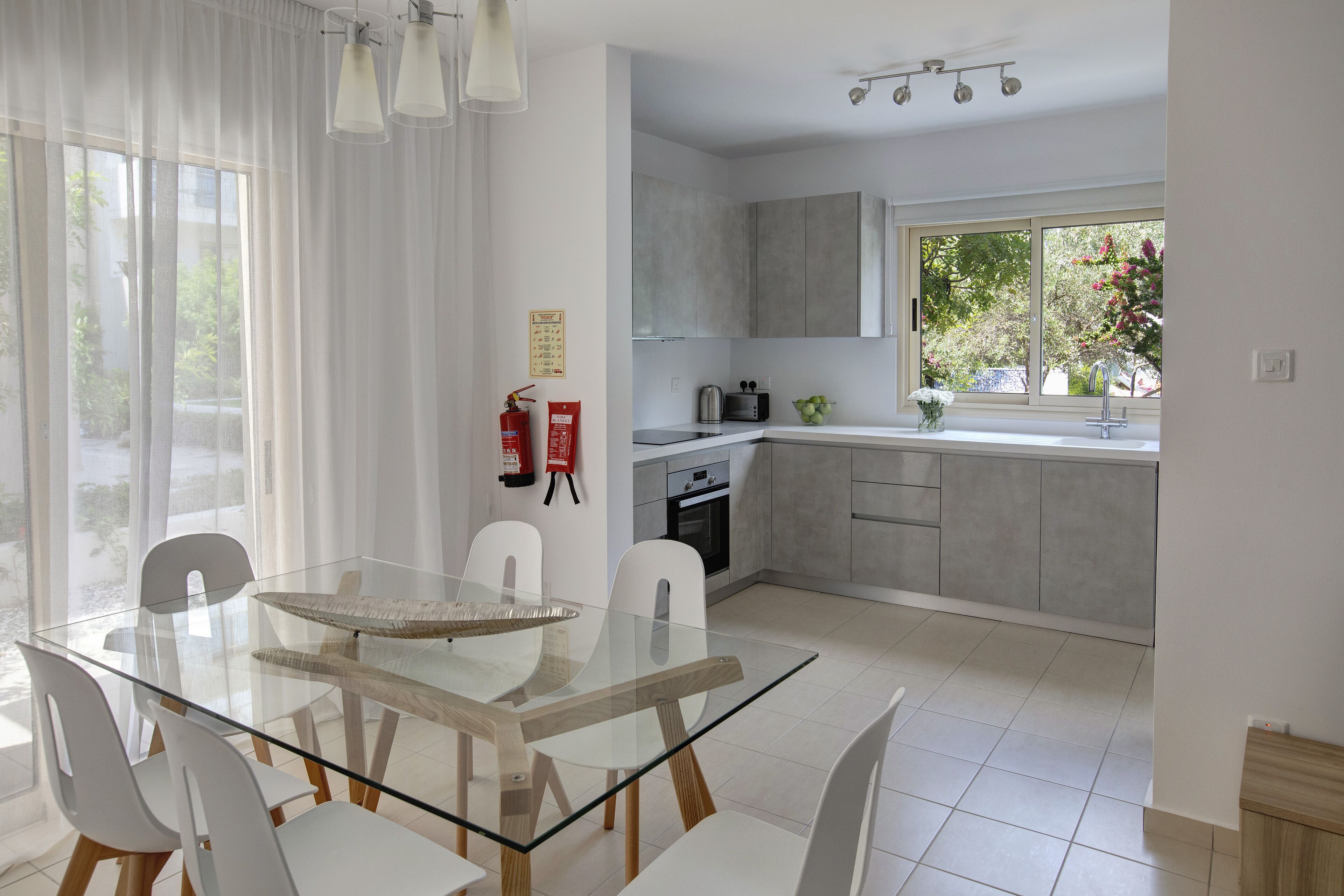 A modern kitchen with a glass dining table, white chairs, and grey cabinets.
