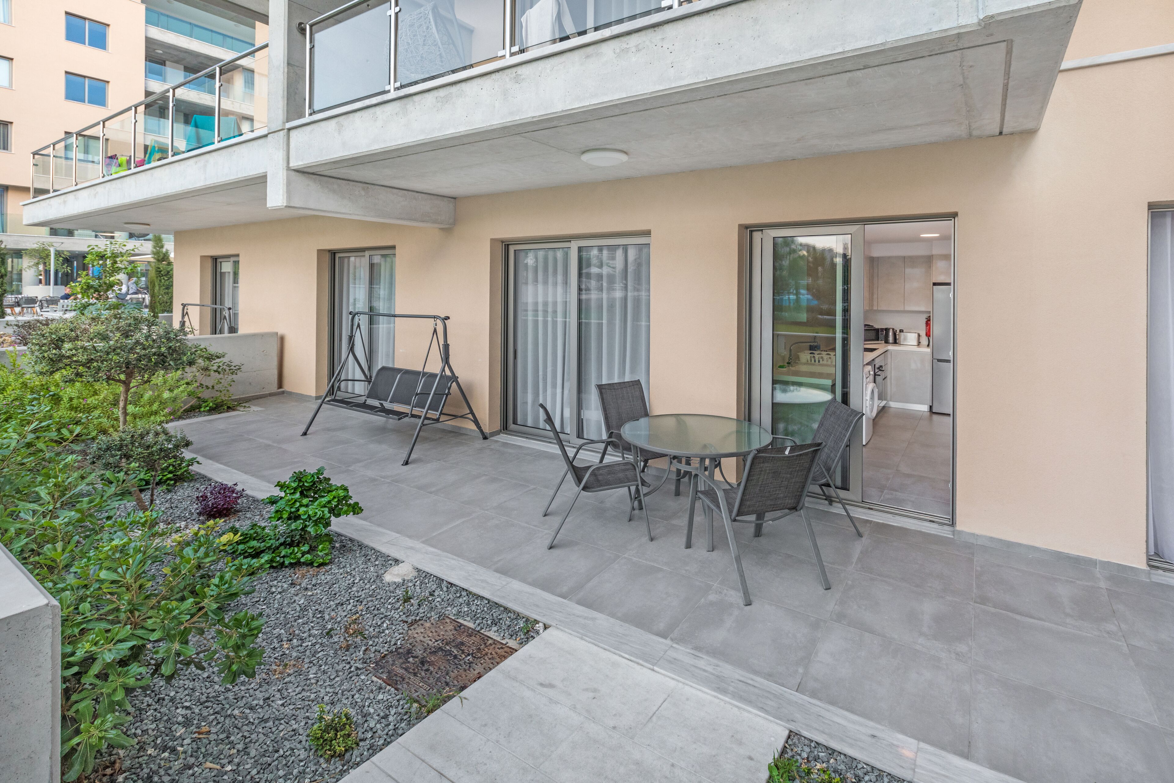 A modern outdoor patio with a glass table and chairs, surrounded by a gravel garden and a concrete pathway.