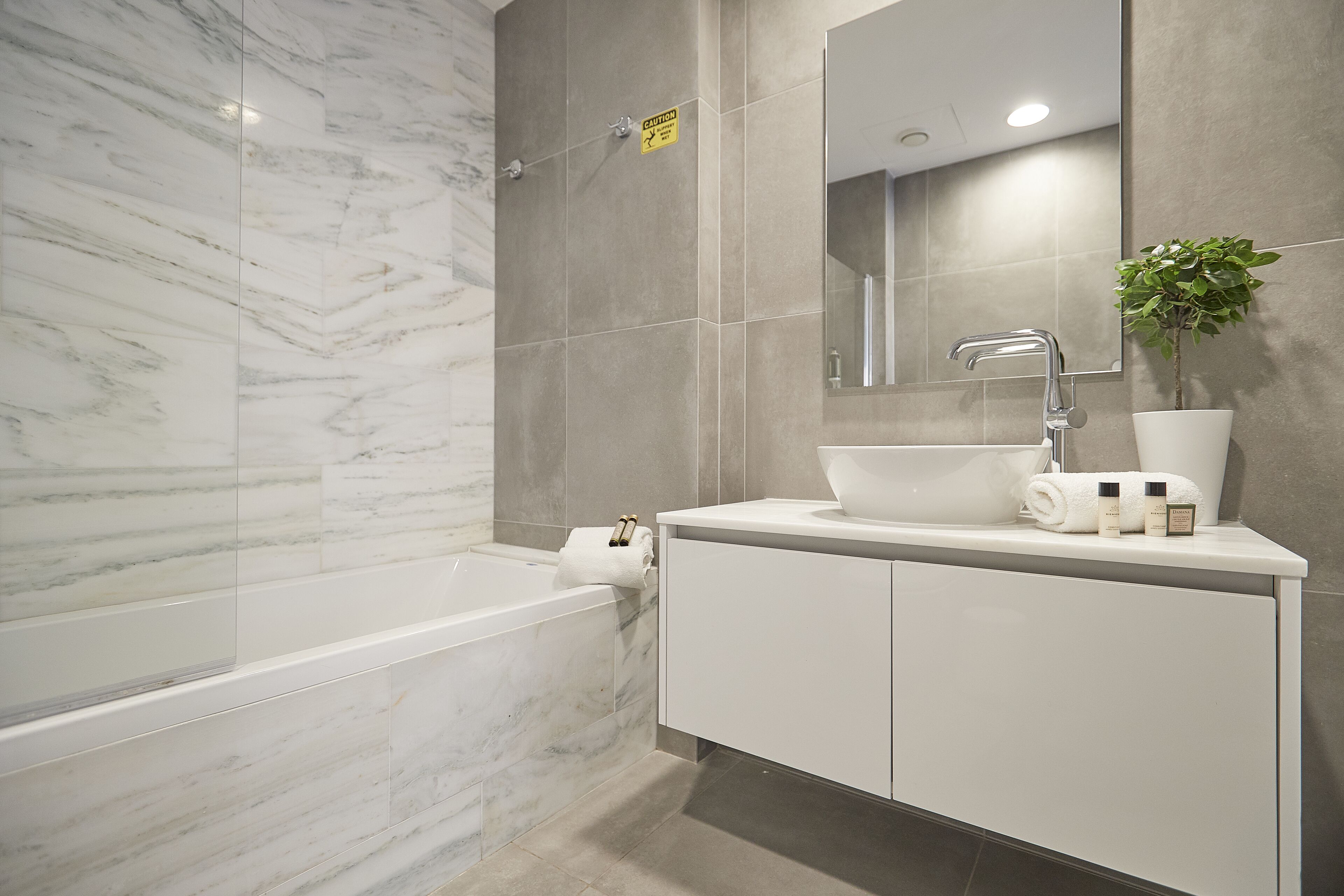 A modern bathroom with a marble wall, a bathtub, a sink with toiletries, and a potted plant.