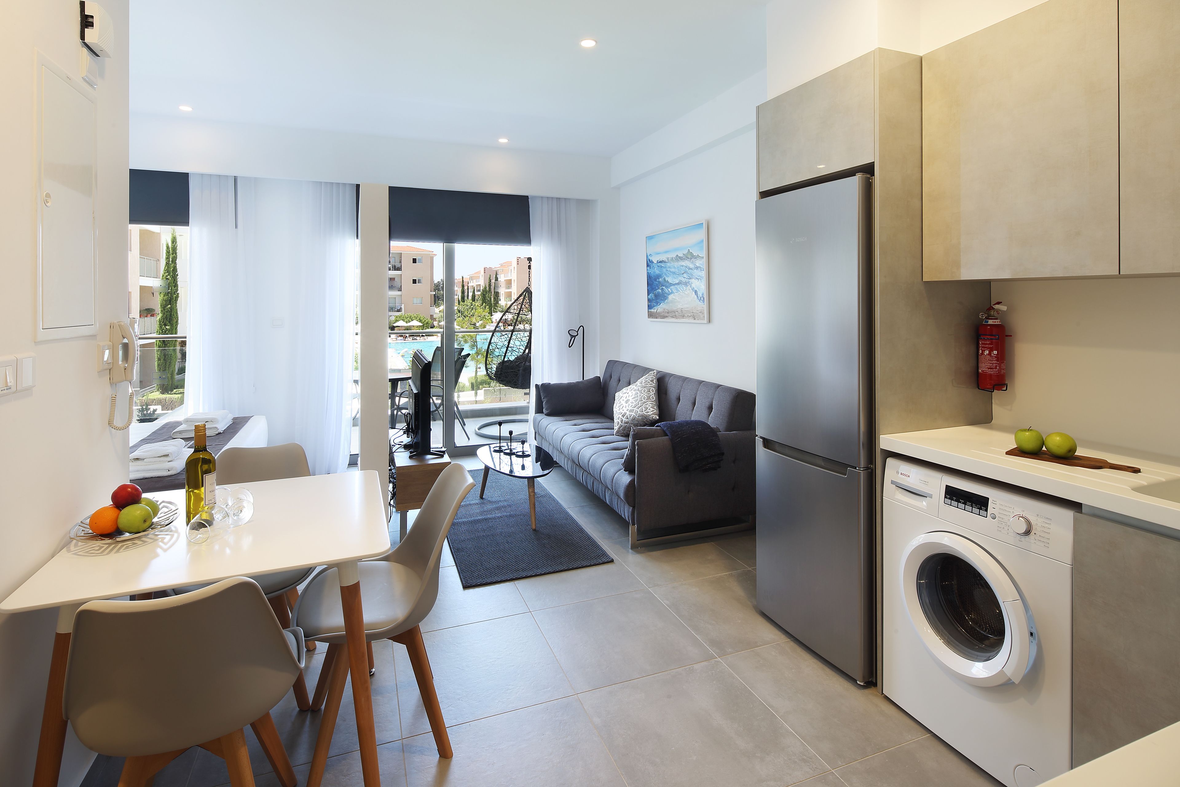 A modern kitchen with a built-in washing machine, a dining area with a table and chairs, a sofa, and a view of a pool and buildings in the background.