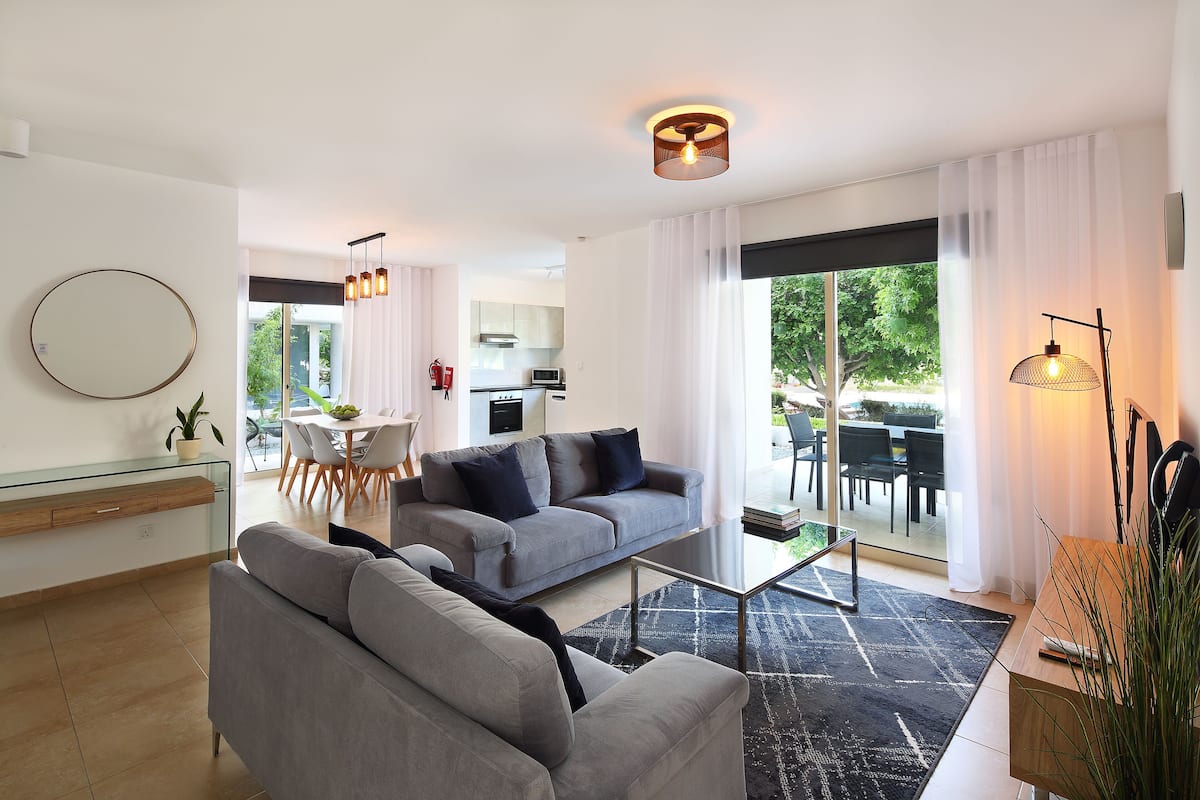 Una sala de estar moderna con un sofá gris, una mesa de centro de vidrio y un comedor con una mesa y sillas blancas.