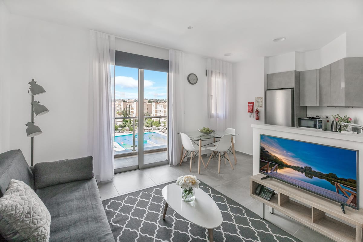 Una sala de estar moderna con un sofá gris, una mesa de centro blanca con flores, un televisor de pantalla plana y un comedor con una mesa y sillas de vidrio.