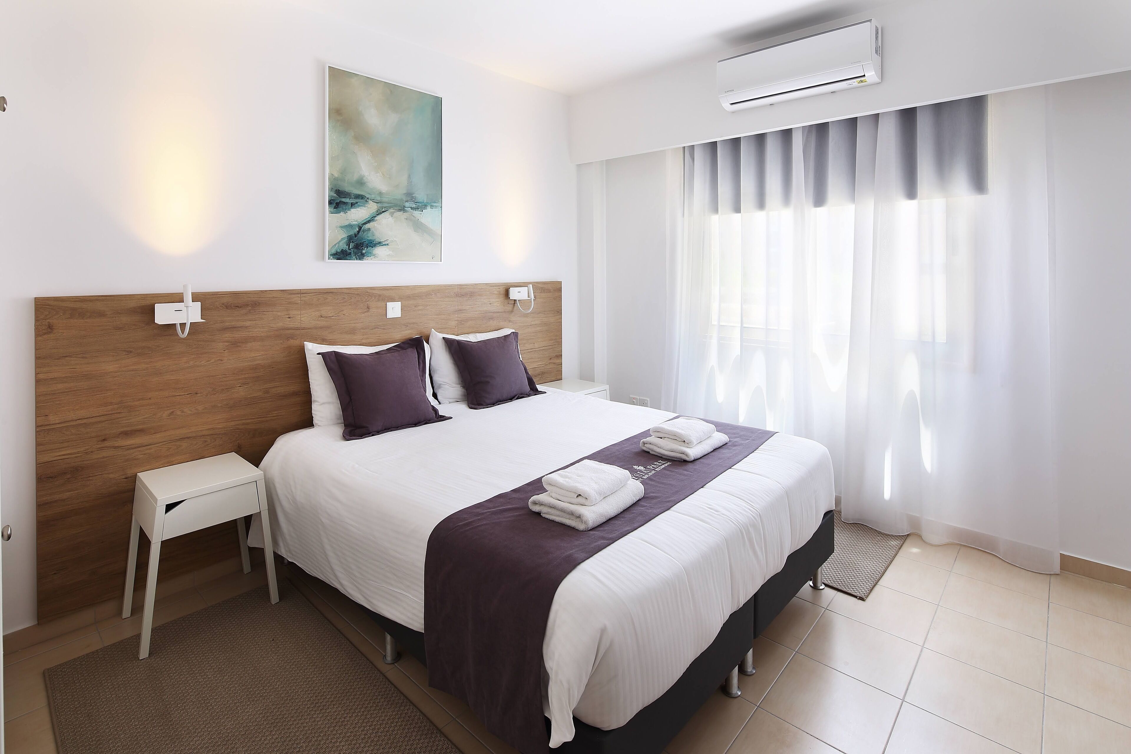 Une chambre d’hôtel moderne avec une tête de lit en bois, un grand lit avec une literie blanche, une petite table de chevet et une grande fenêtre avec des rideaux légers.