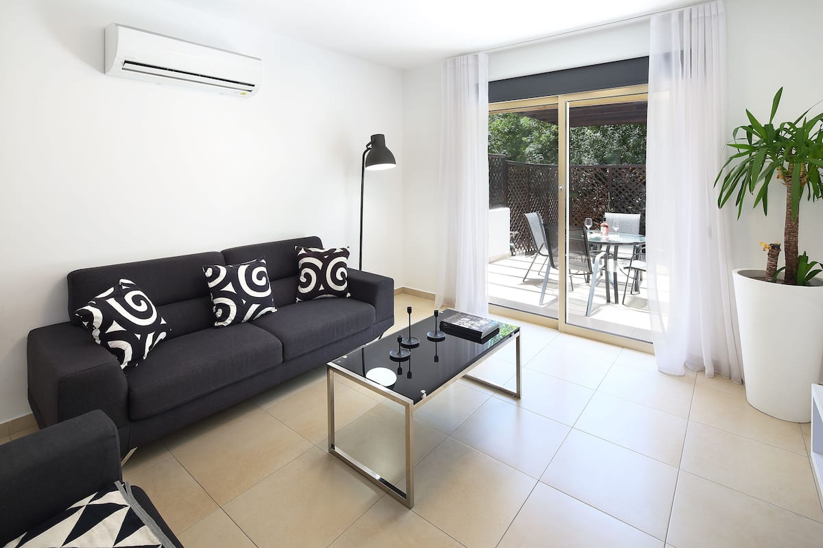 Una sala de estar moderna con un sofá negro, una mesa de centro de vidrio y un ventanal con vistas a un patio exterior.