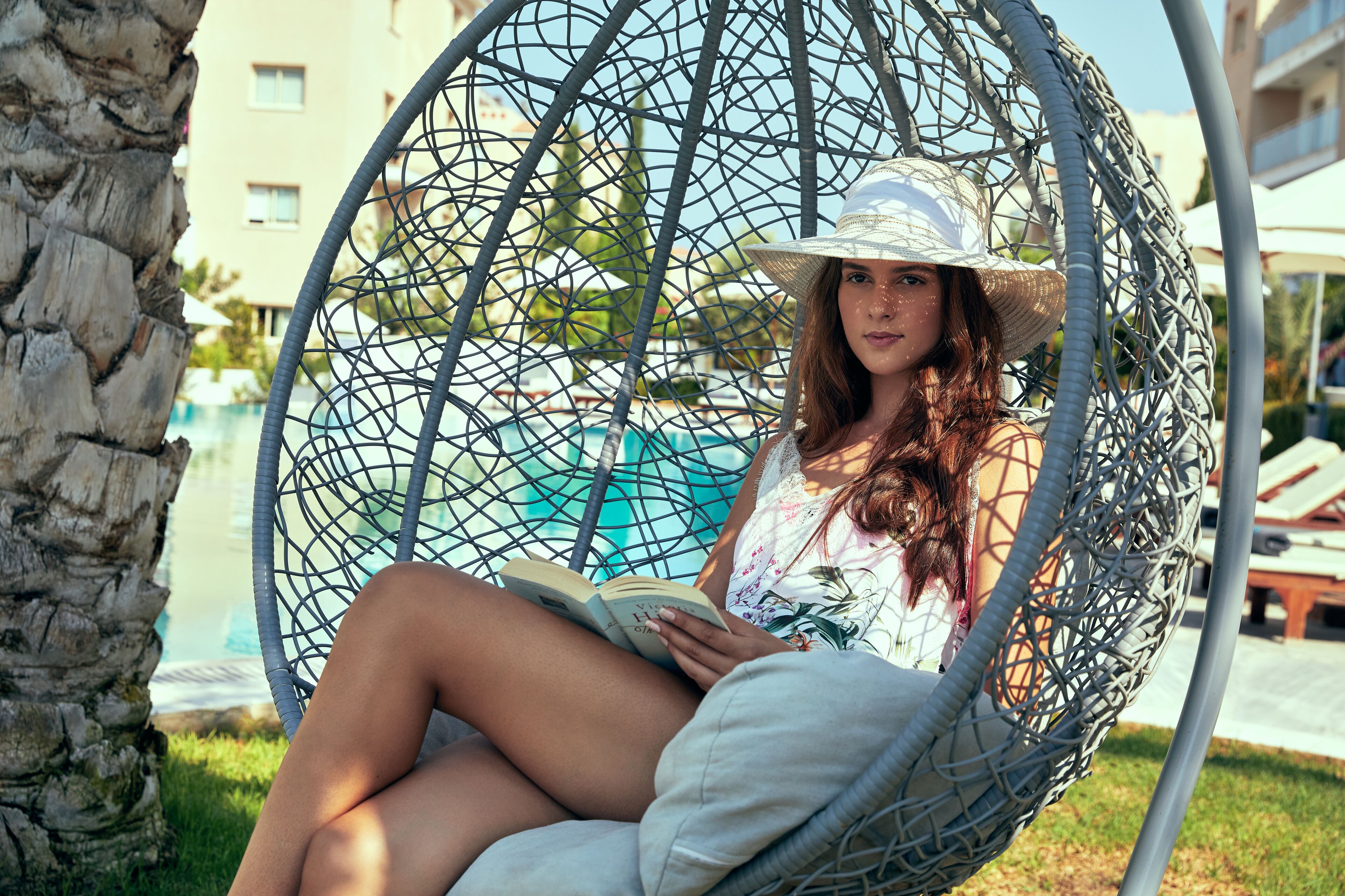 A person sitting in a wicker swing chair by a pool, reading a book.