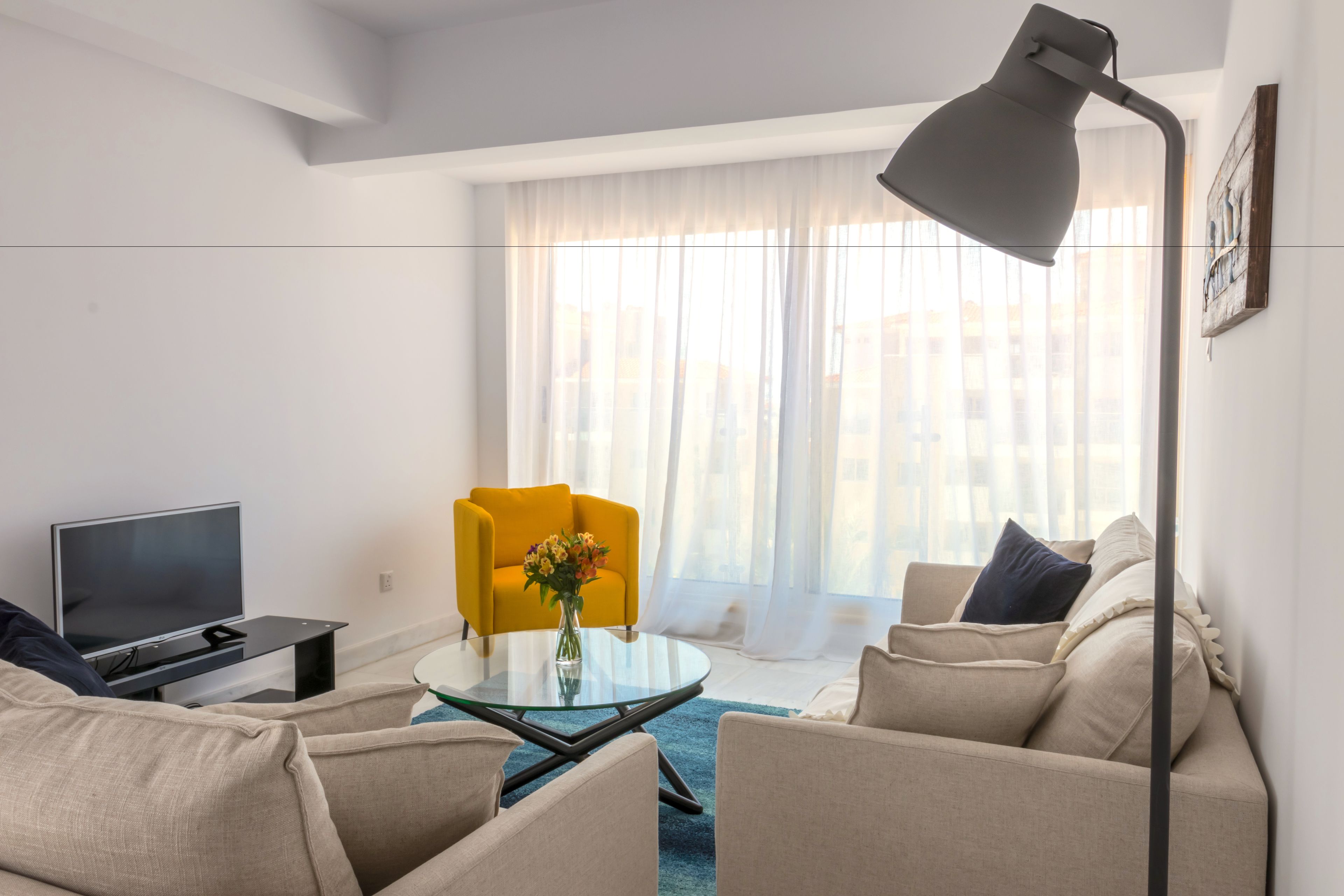 A modern living room with a sofa, a yellow armchair, a glass coffee table, and a TV on a stand.